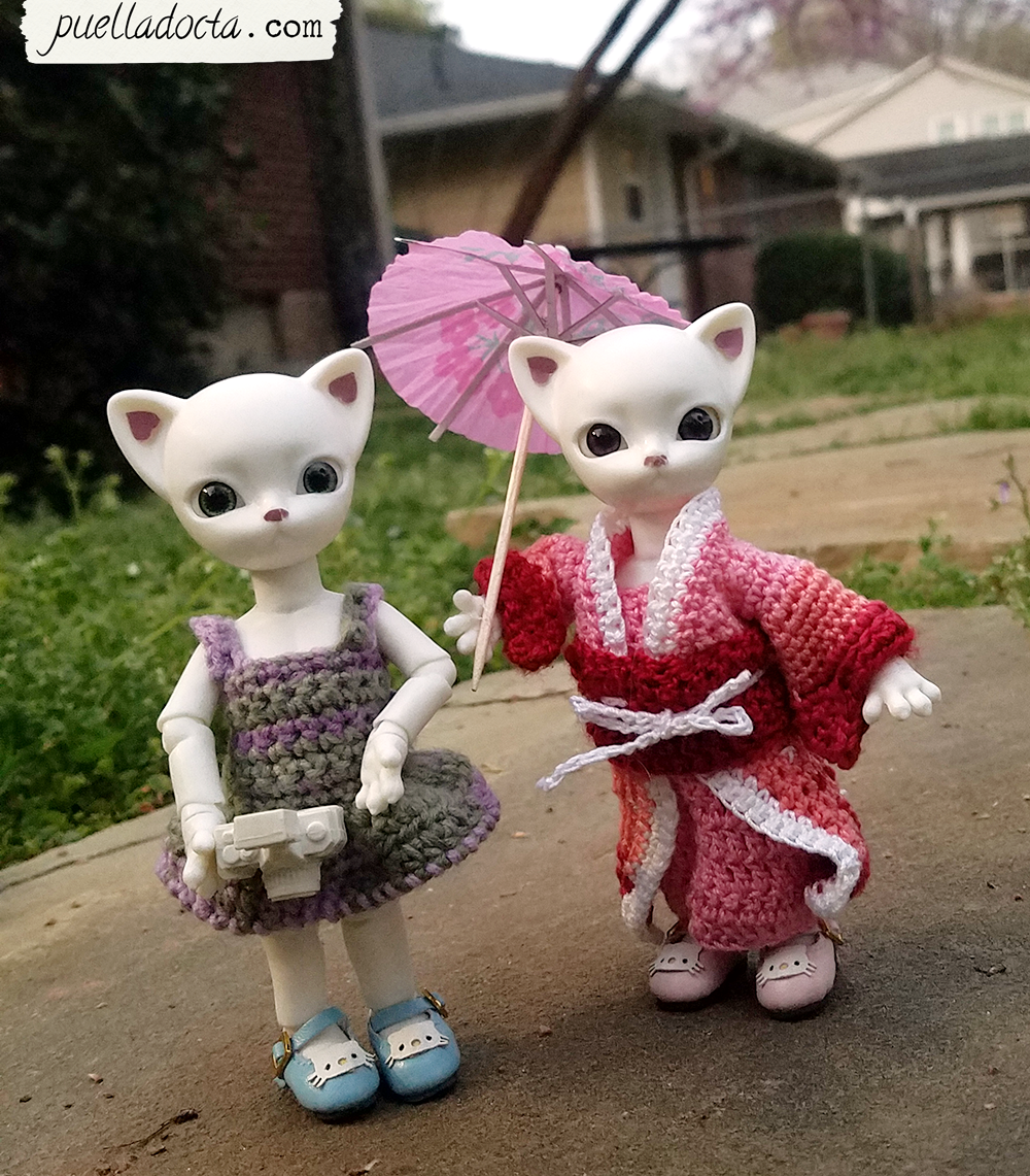 Two white ball jointed cat dolls, one wearing a green and purple crocheted dress with straps, one in a crocheted pink and red kimono.